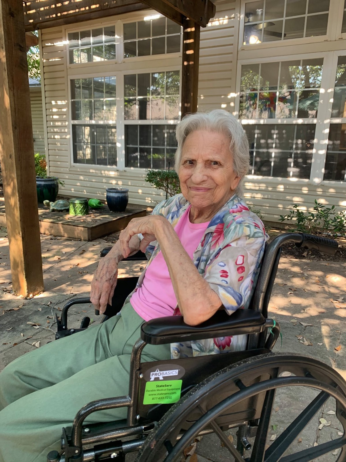 An elderly woman sitting in a wheelchair outdoors, surrounded by garden plants.