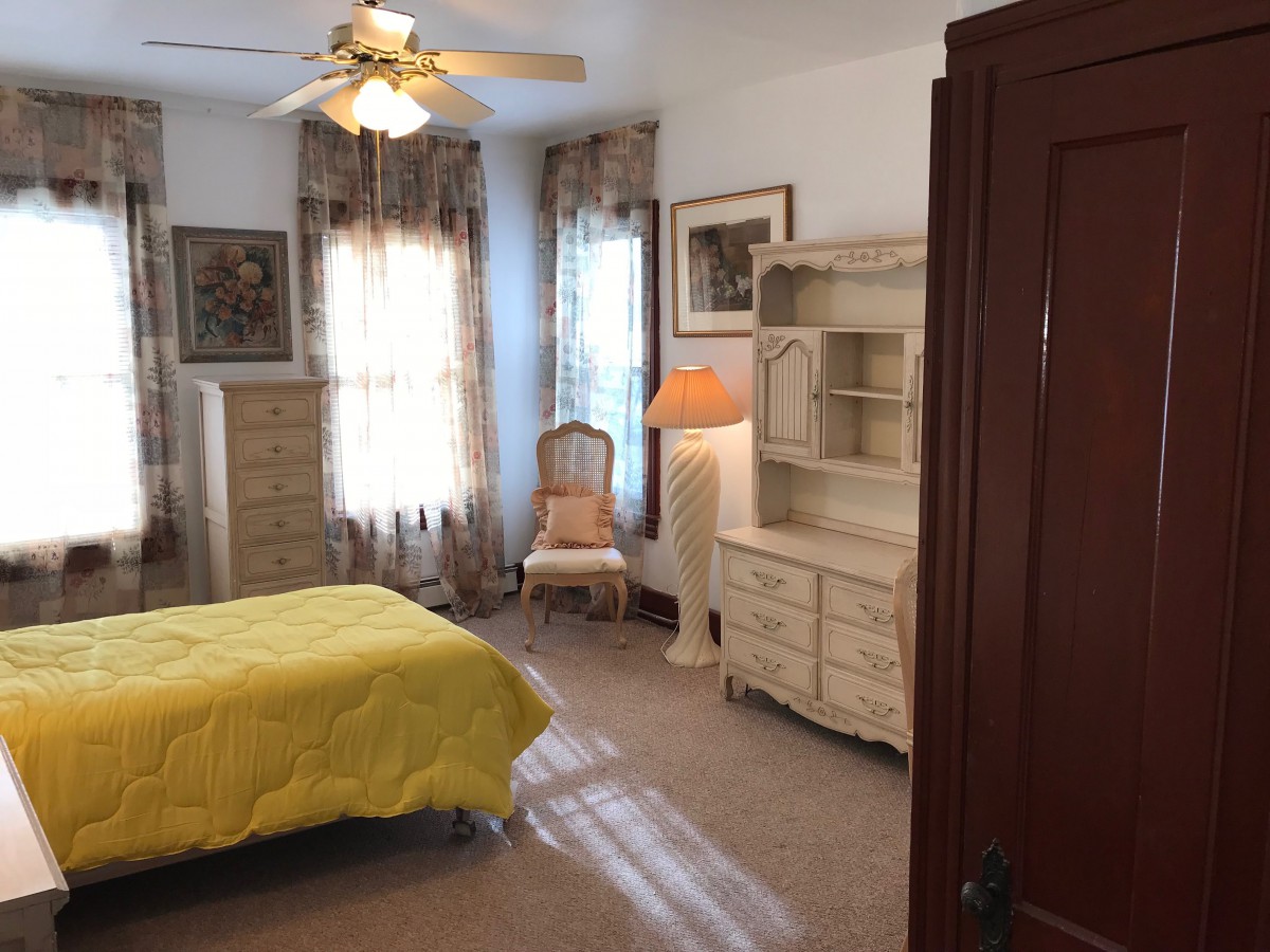 A cozy bedroom with vintage-style furniture, featuring a bed with a yellow cover, a dresser, and natural light streaming through the windows.
