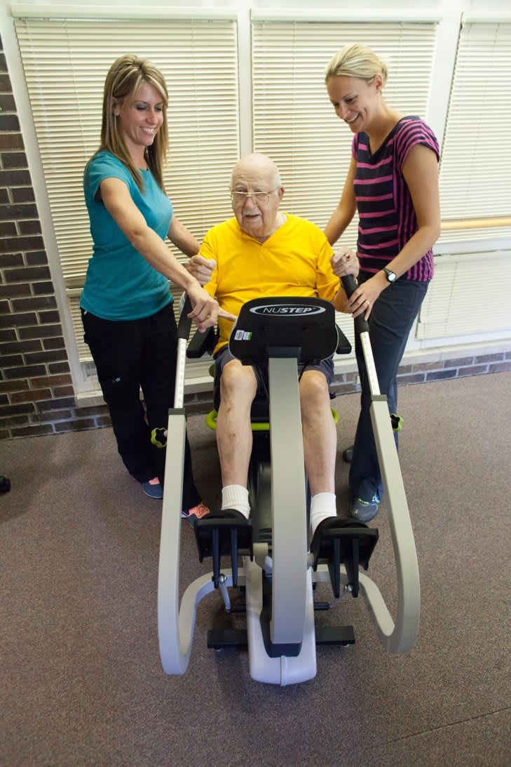 An elderly man receiving assistance with physical therapy exercises on a specialized machine.