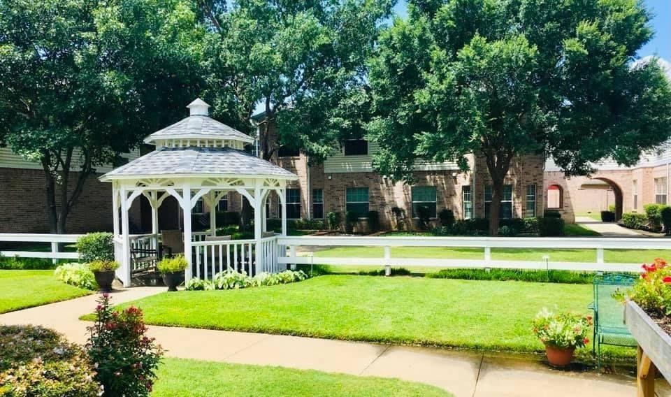 A beautiful garden with a white gazebo, surrounded by lush green grass and trees, adjacent to a building.