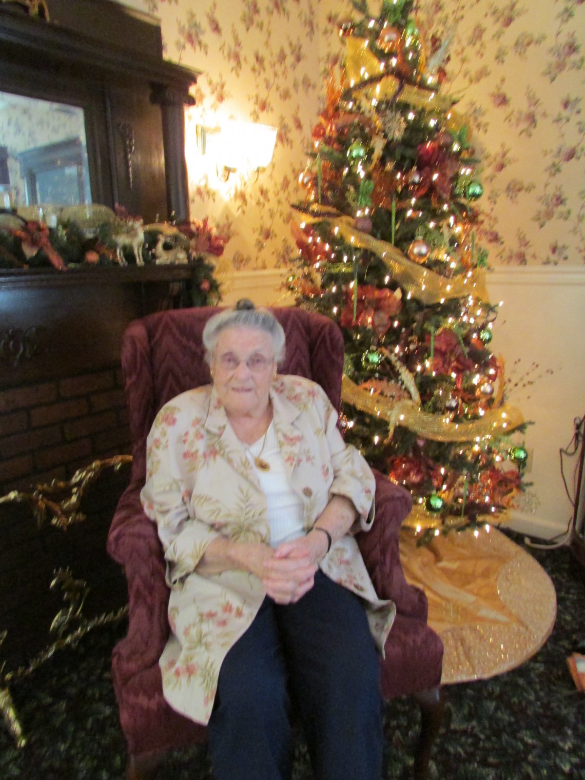 An elderly woman sitting in an armchair next to a decorated Christmas tree.