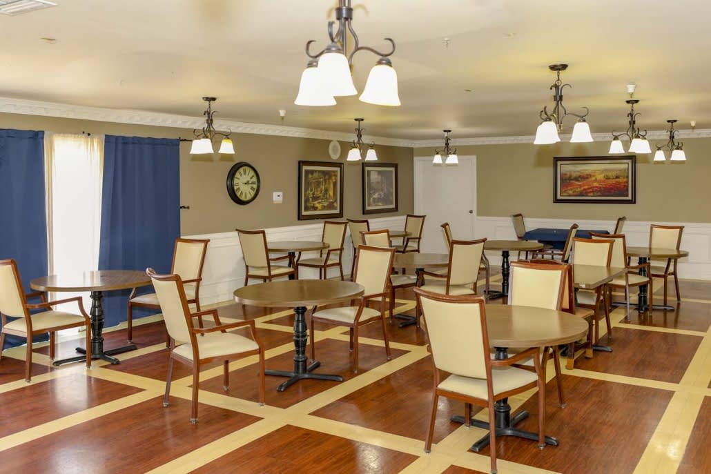 A well-lit dining room with several tables and chairs, featuring decorative paintings on the walls and a wooden floor.