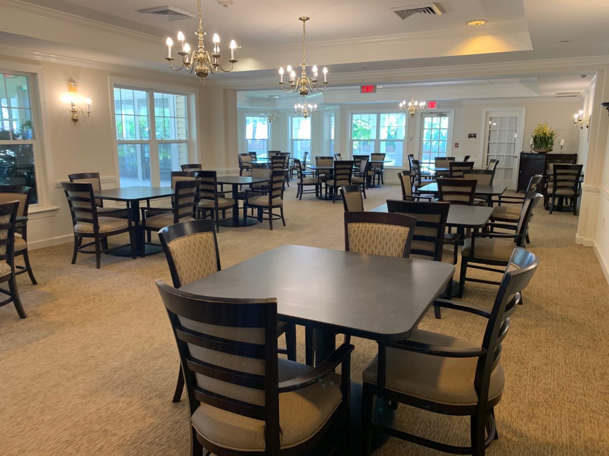 A spacious dining room with multiple tables and chairs, featuring elegant chandeliers and large windows.