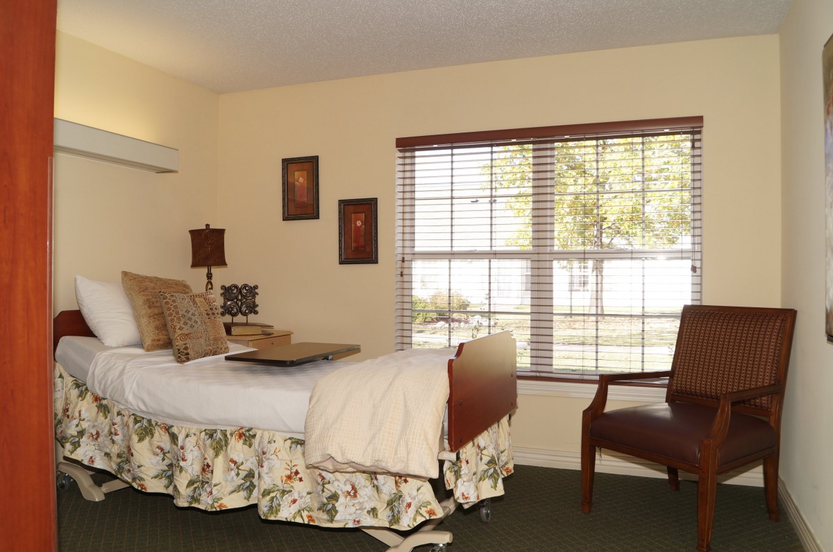 A cozy bedroom interior featuring a neatly made bed with floral bedding, a wooden chair, and a large window with blinds.