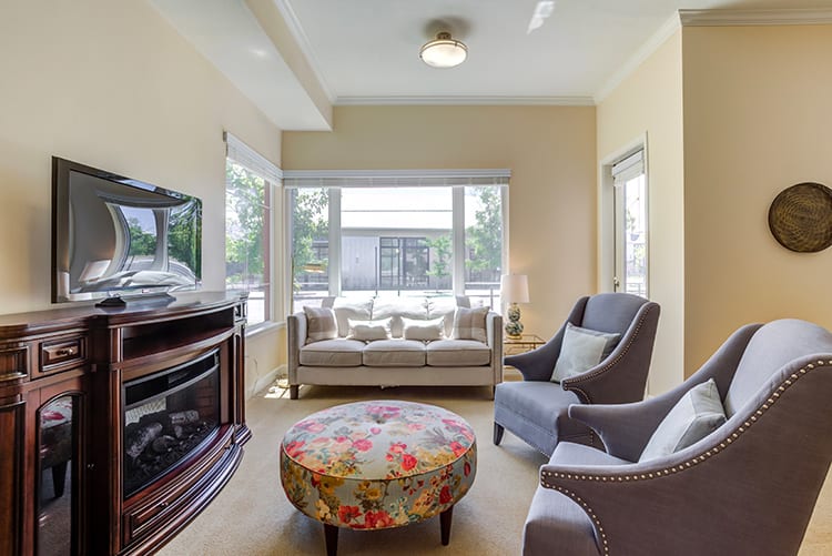 A cozy living room with a sofa, armchairs, a floral ottoman, and large windows providing natural light.