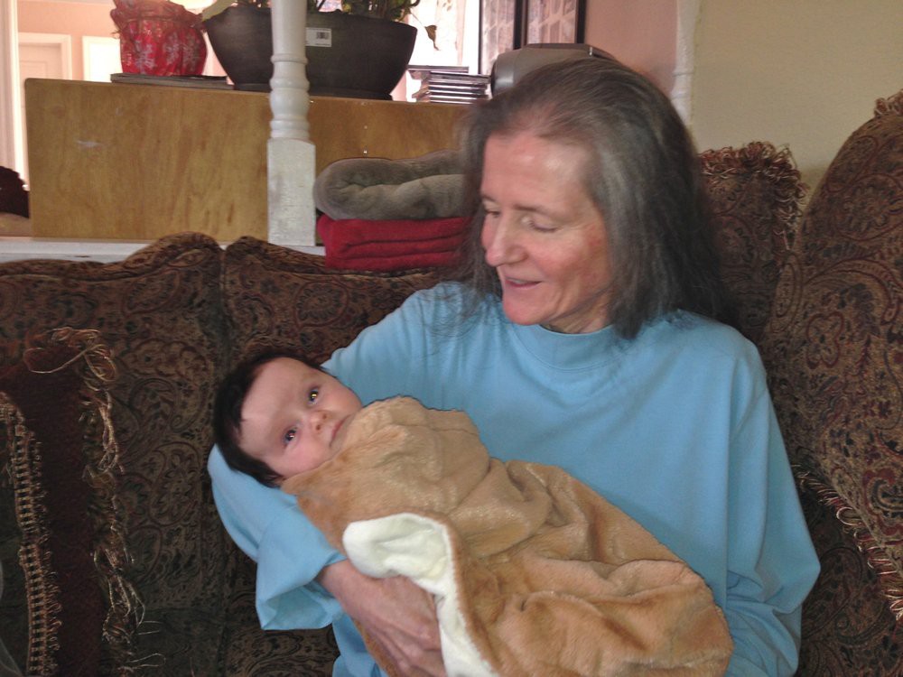An elderly woman smiling and holding a baby wrapped in a blanket on a couch.