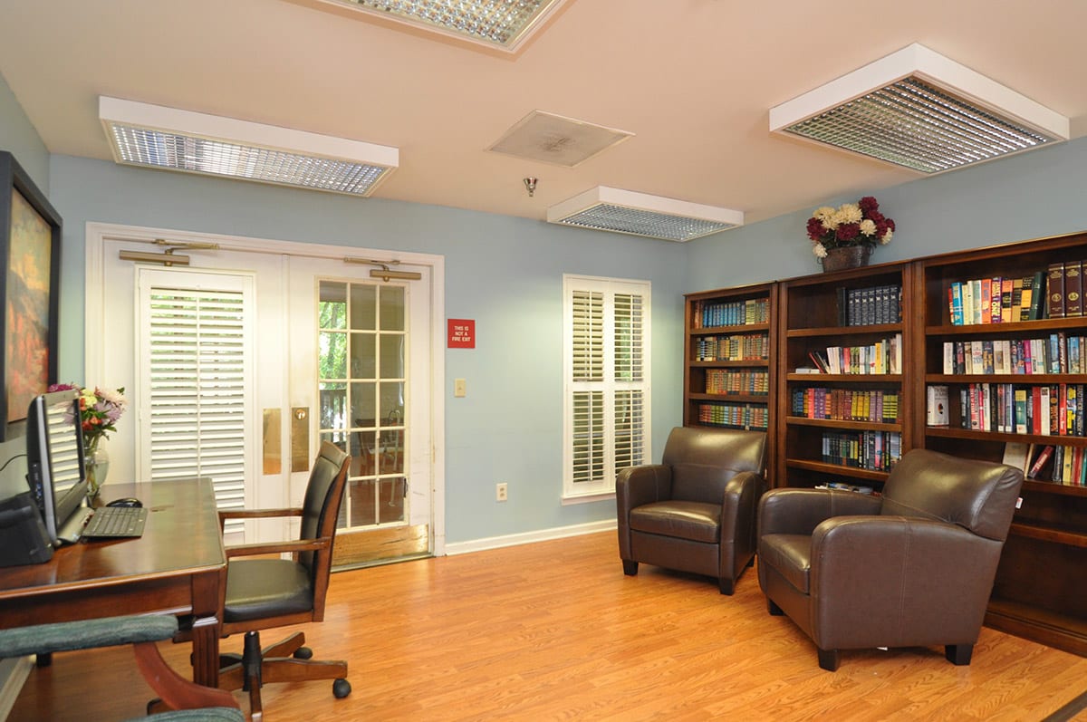 A cozy library room featuring bookshelves, comfortable armchairs, and a wooden table with office chairs.
