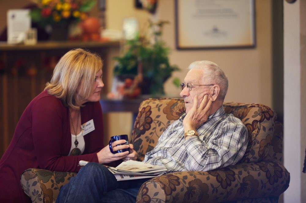 A caregiver engages in a warm conversation with an elderly man in a comfortable indoor setting.
