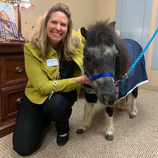A woman in a green jacket smiles while posing with a small pony indoors.