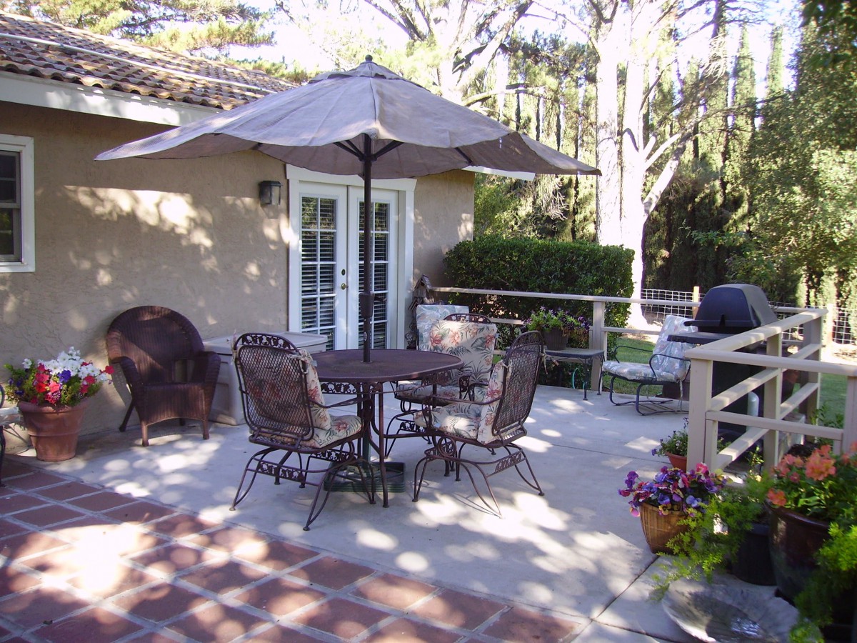 A sunny patio with outdoor furniture, including a table and chairs under an umbrella, surrounded by greenery.