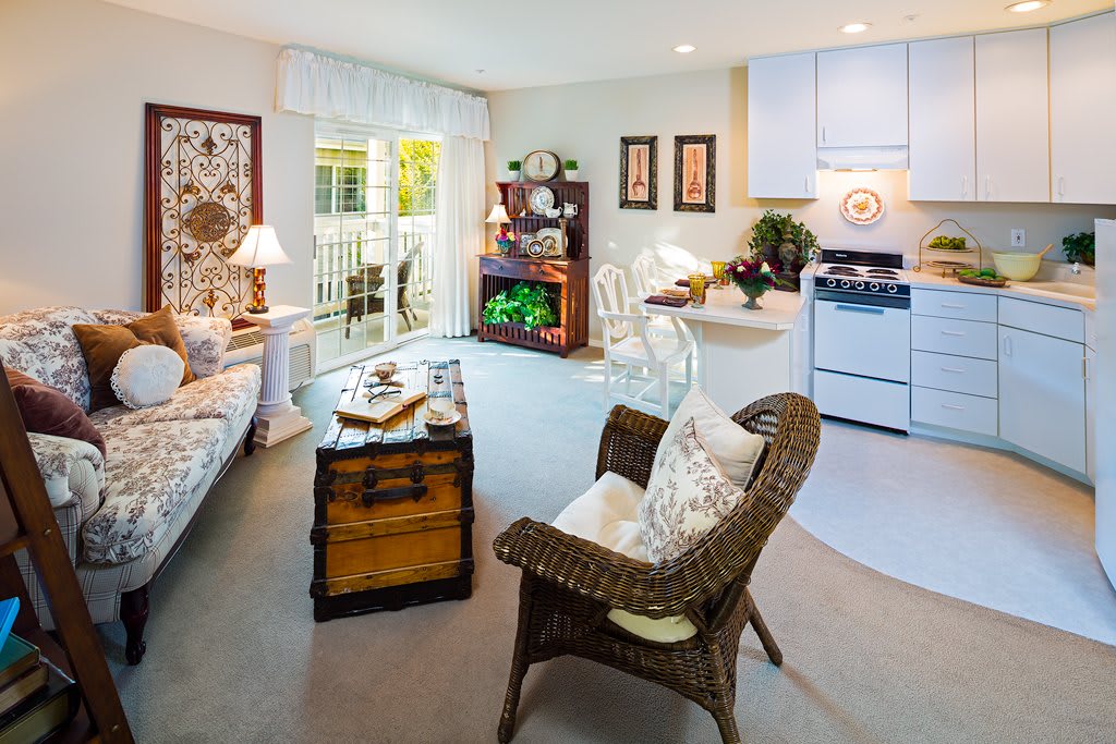 A cozy living room with a rattan chair, wooden chest table, and a bright ambiance connecting to a small kitchenette.