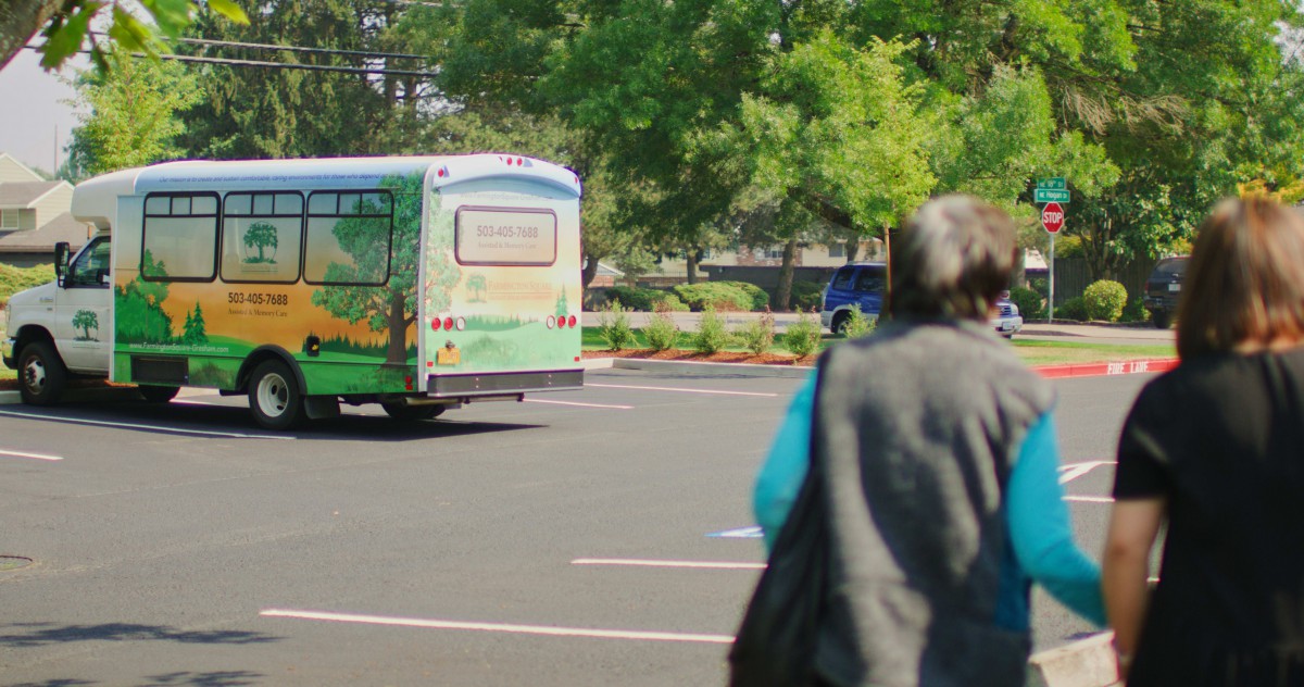 A person walking in a parking lot towards a parked bus with trees in the background.