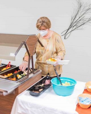 A person wearing a mask serves themselves food from a buffet table.