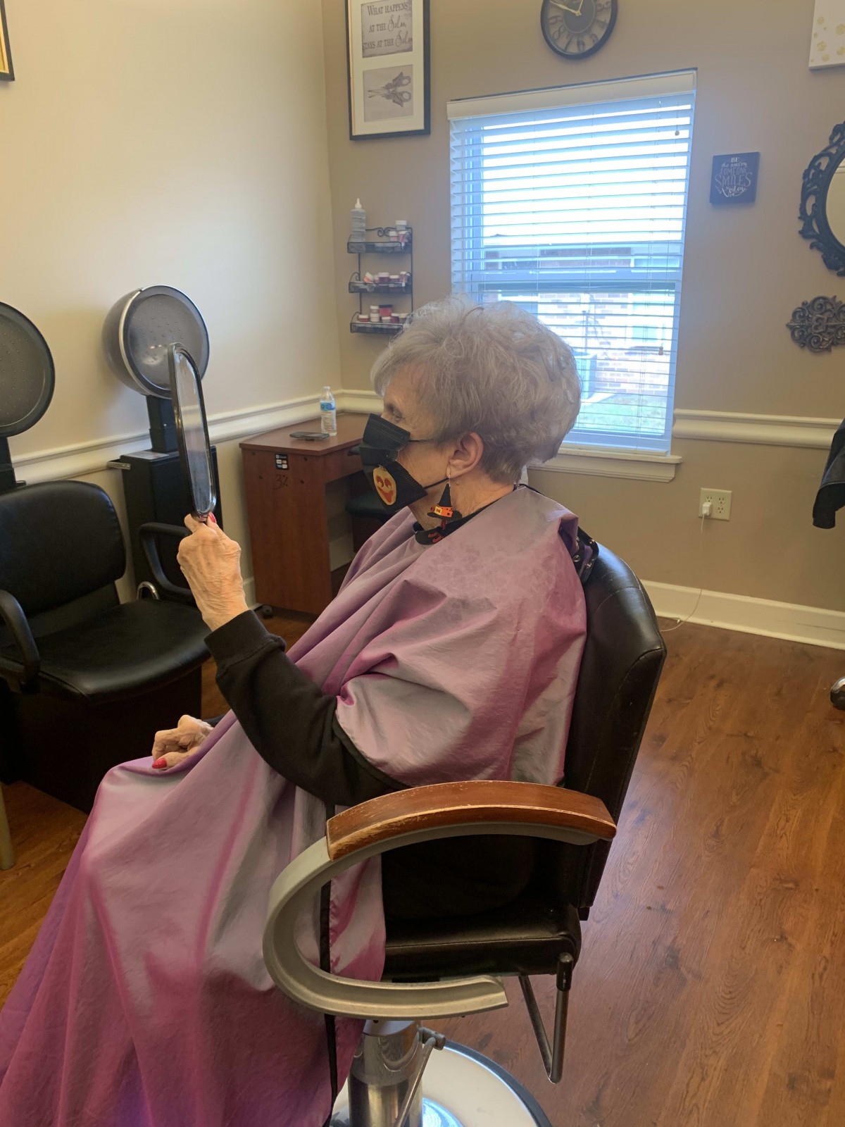 A senior woman wearing a cape looks at her reflection in a hand mirror inside a hair salon.