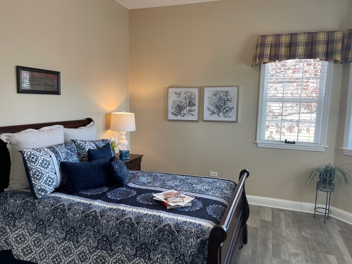 A cozy bedroom featuring a patterned bedspread, two framed artworks above the bed, a bedside lamp, and a window with plaid curtains.