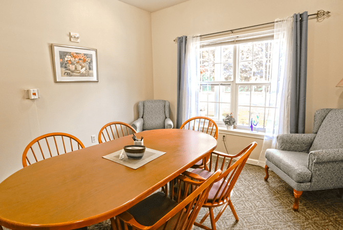 A cozy dining room with a wooden table and chairs, featuring a large window and an armchair.