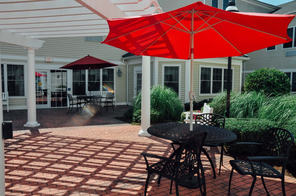 A sunny patio with red umbrellas, providing shade over tables and chairs on a brick floor.