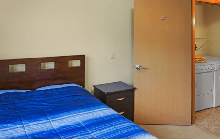 A simple bedroom with a bed, blue blanket, nightstand, and door.