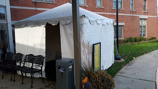 A white tent set up in a public area, featuring a chair and a nearby trash bin.
