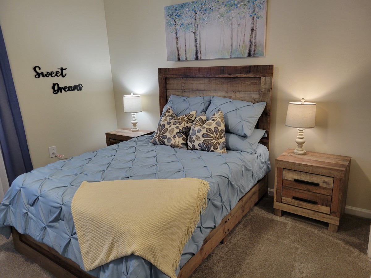 A cozy bedroom featuring a wooden bed with blue bedding and decorative pillows, flanked by matching nightstands and lamps, complemented by wall art.