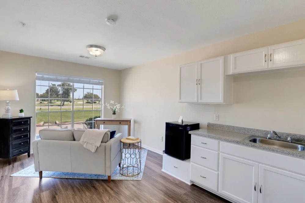 A modern and minimalist living room with an adjacent kitchenette, featuring a large window and wooden flooring.