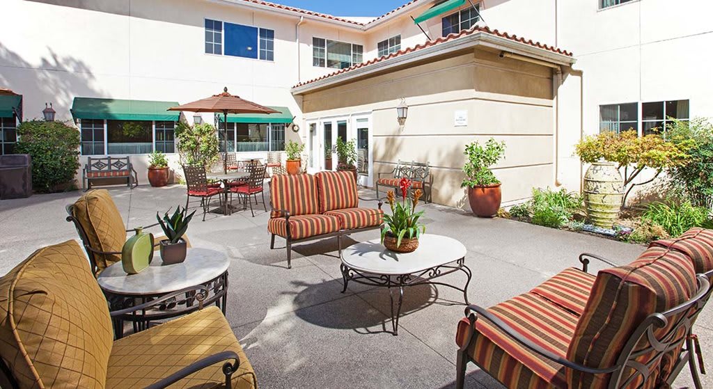 A cozy outdoor patio with comfortable seating and tables, surrounded by potted plants against the backdrop of a building exterior.