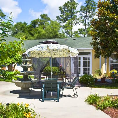 A charming garden patio with a table and chairs under an umbrella, surrounded by greenery and featuring a central fountain on a sunny day.