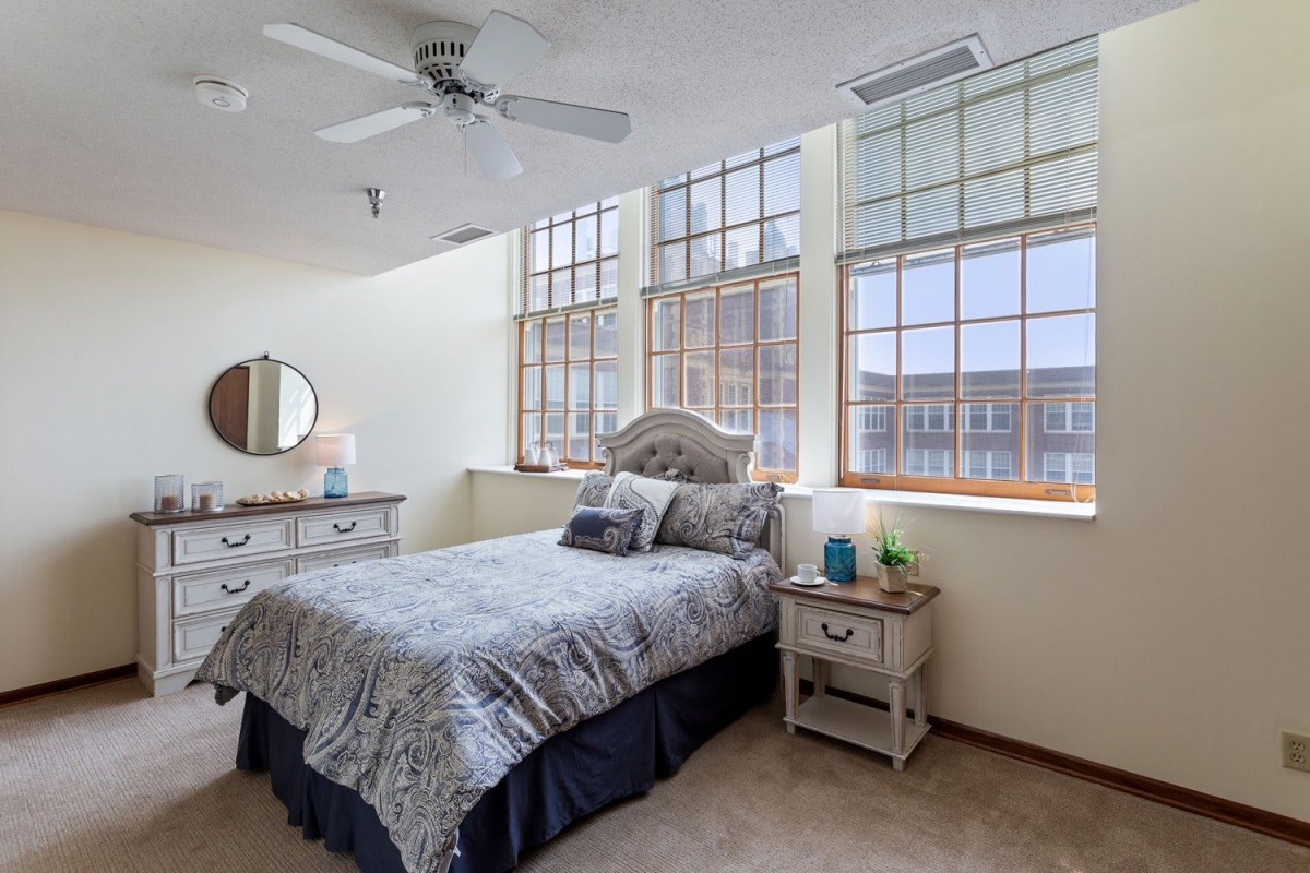 A cozy bedroom featuring a bed with patterned bedding, large windows allowing natural light, and a ceiling fan.