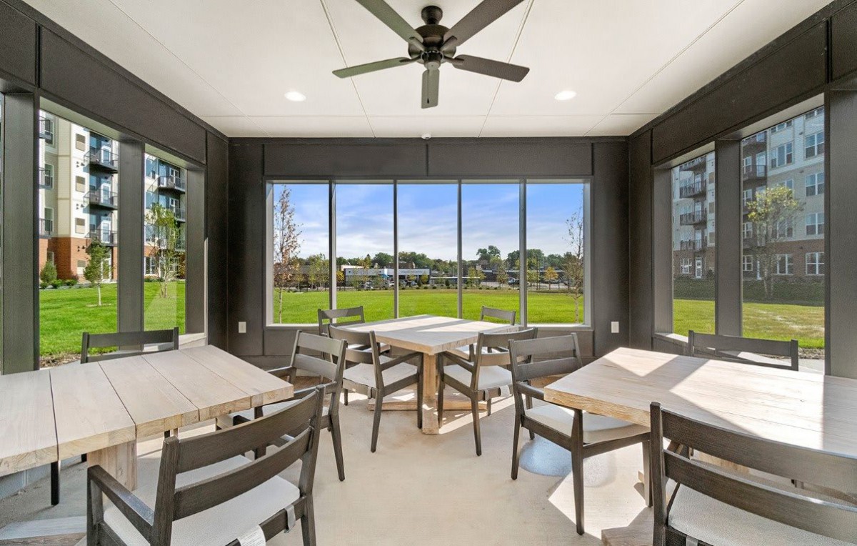 A bright dining area with multiple tables and chairs, featuring large windows providing a view of the outdoors.