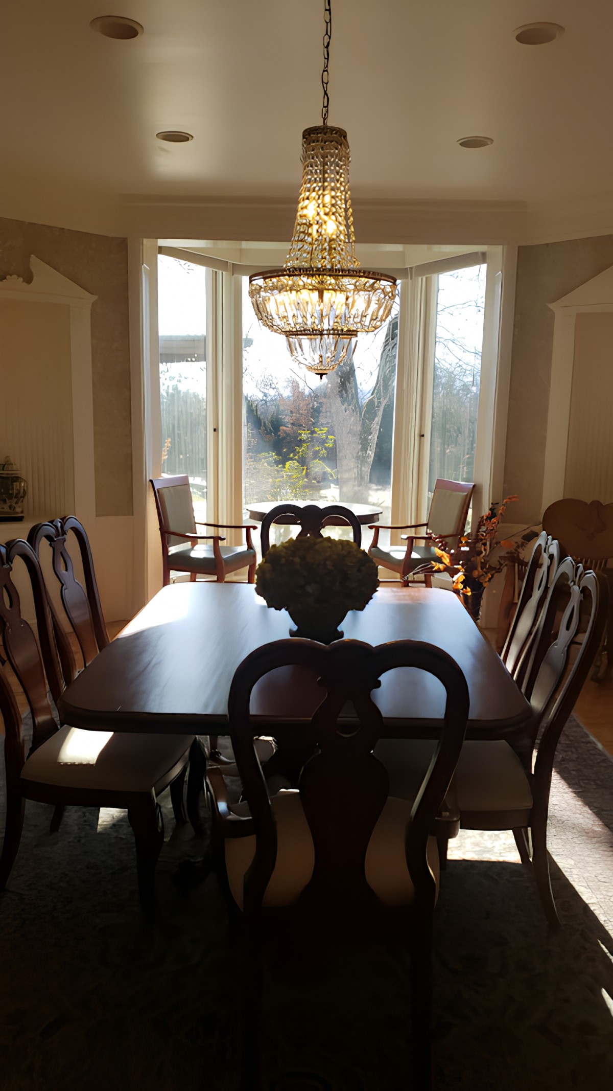 A dining room with a wooden table and chairs, illuminated by a chandelier, showcasing a bay window with natural light.