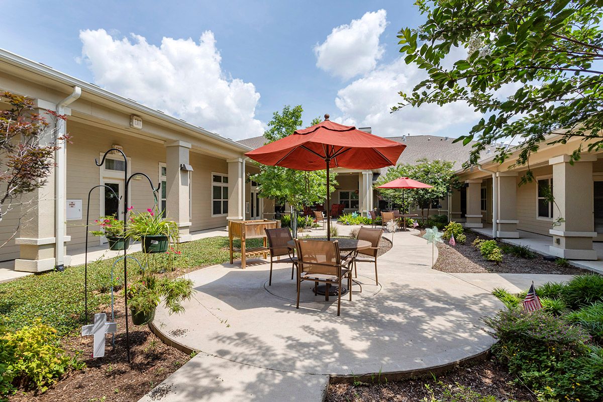 A sunny outdoor patio area with tables, chairs, and red umbrellas surrounded by landscaped gardens.