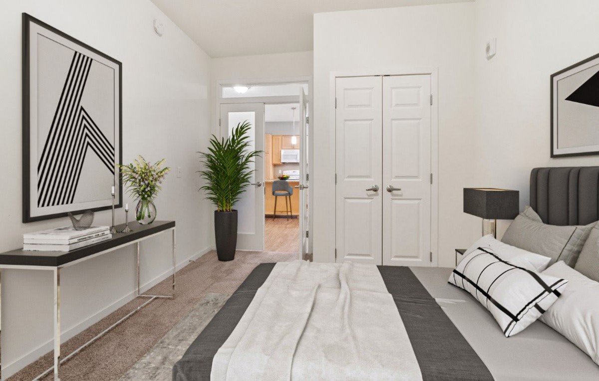 A modern, minimalist bedroom with white walls and a carpeted floor, featuring a bed, decorative artwork, and a green plant. The room has double doors leading to another area.