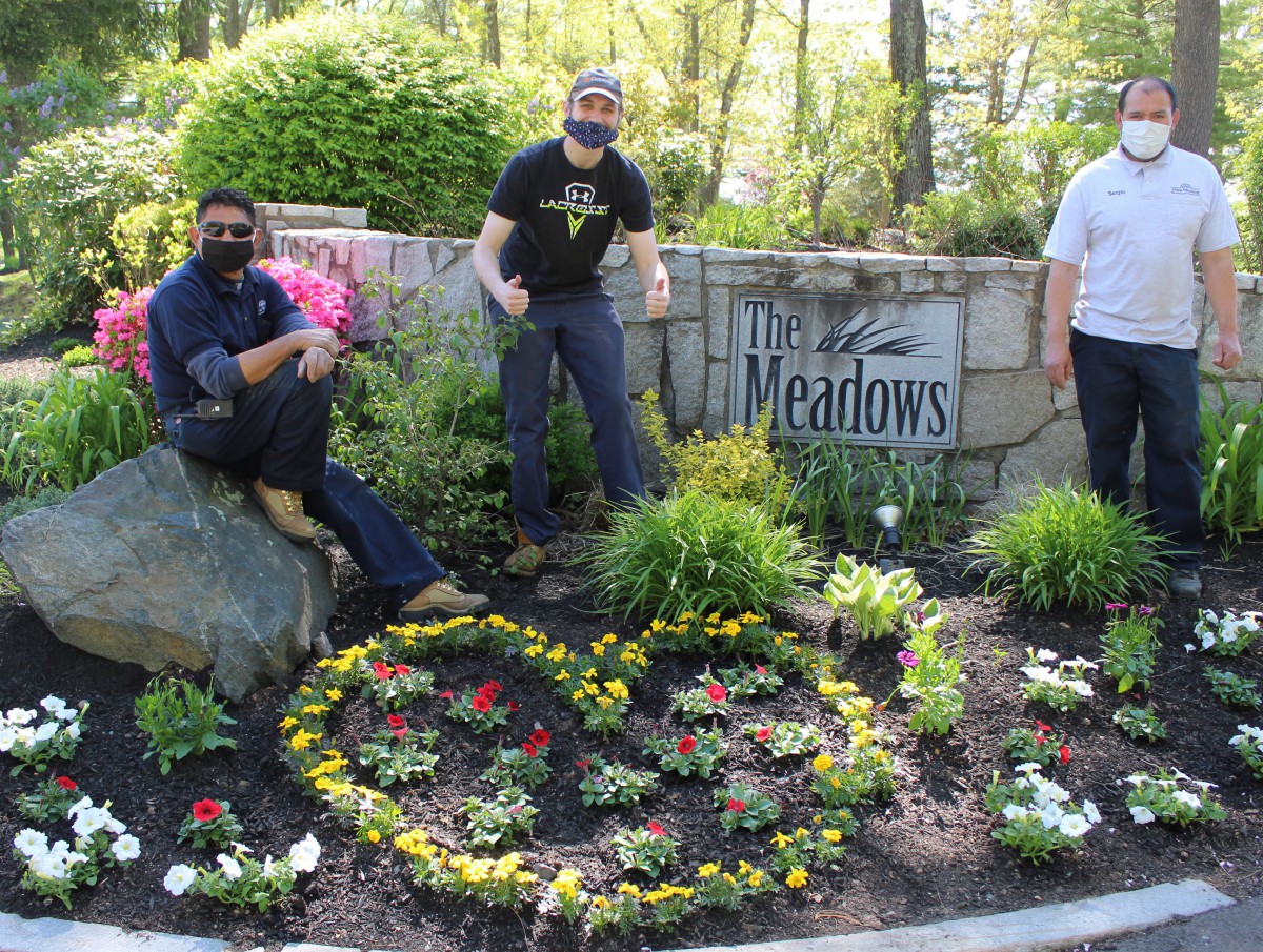 Three individuals posing in a garden setting with colorful flowers and a sign reading 'The Meadows' in the background.