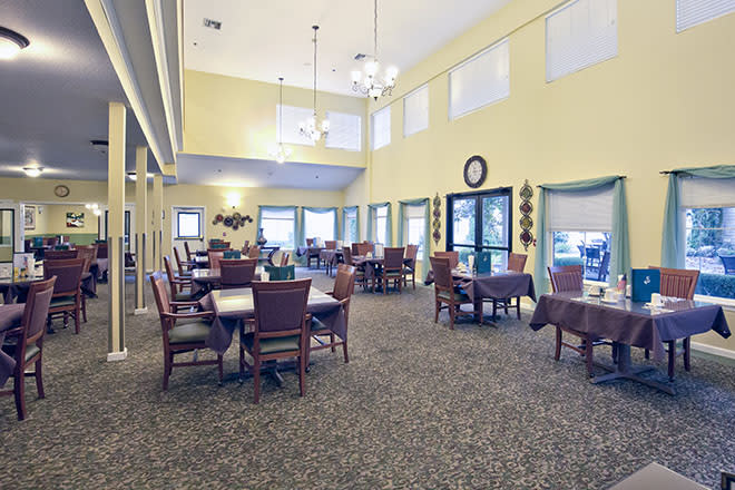 A spacious dining room with tables and chairs, illuminated by chandeliers and natural light from large windows.