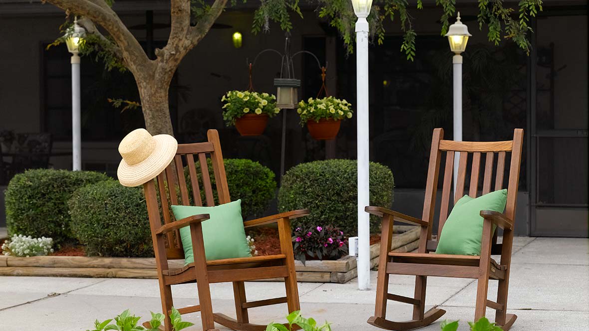 A peaceful patio setting with wooden rocking chairs, green pillows, and a hat placed on one of the chairs surrounded by lush plants.
