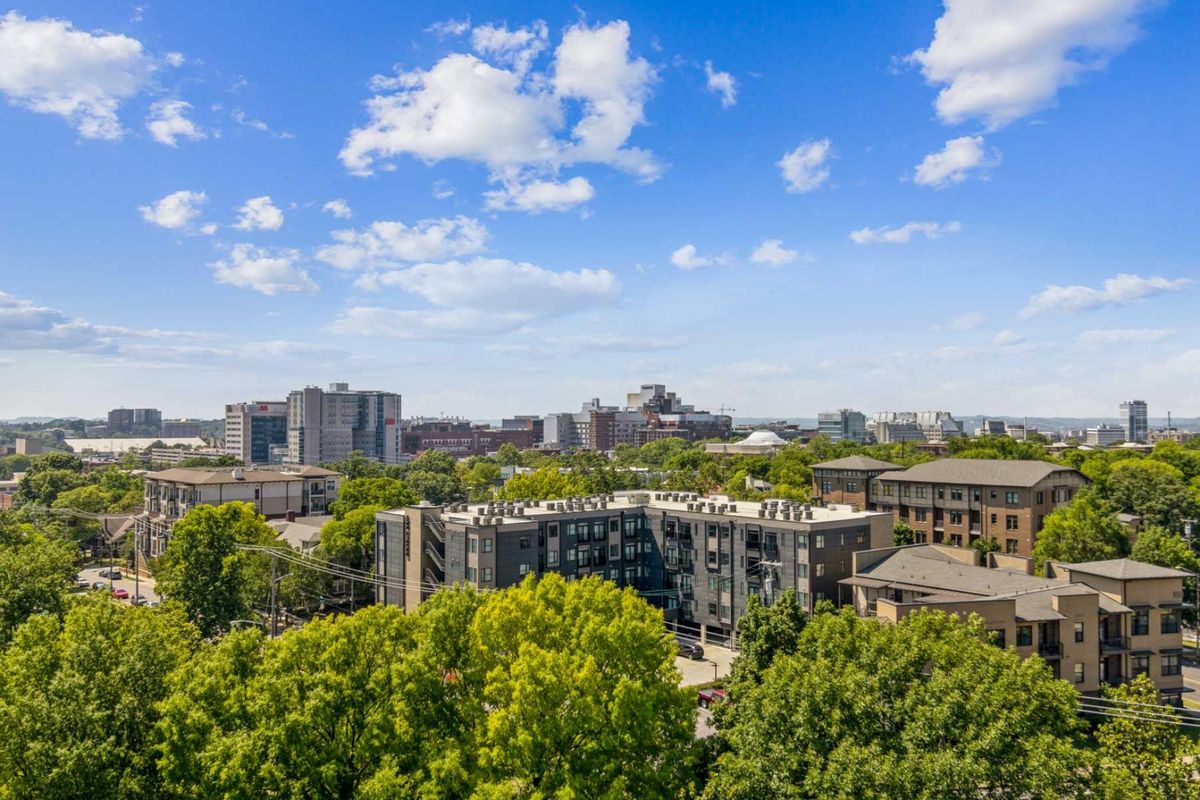 A vibrant urban cityscape with buildings surrounded by lush green trees under a bright blue sky with scattered clouds.