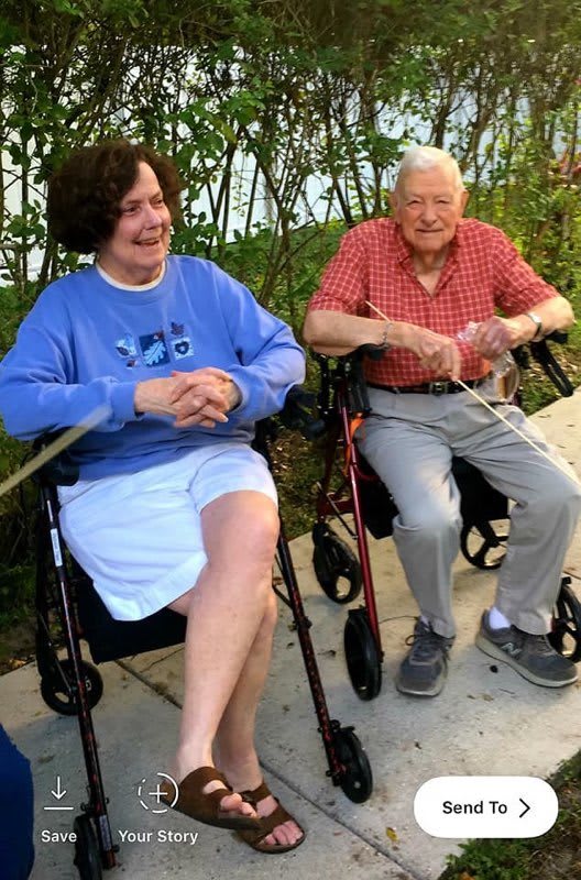 Two elderly individuals sitting outdoors in wheelchairs, enjoying a moment together.