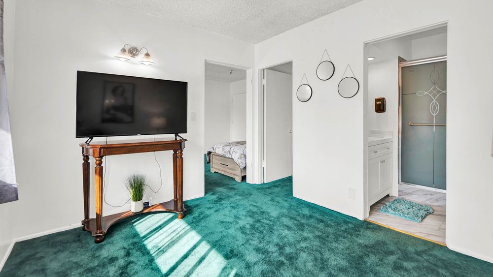 A cozy living area with a green carpet, featuring a large television and wall decor. A glimpse of the bedroom is visible through an open doorway.