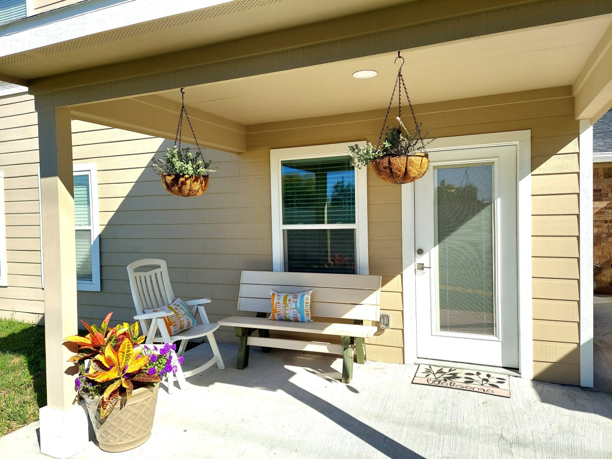 A cozy porch with a bench, chair, and hanging plants on a sunny day.
