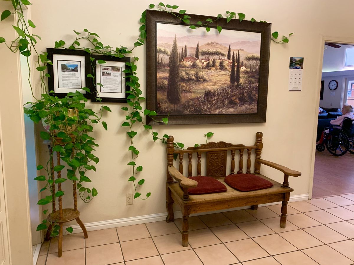 A cozy corner with a wooden bench, adorned with red cushions, framed by leafy green vines and wall art depicting a serene landscape.