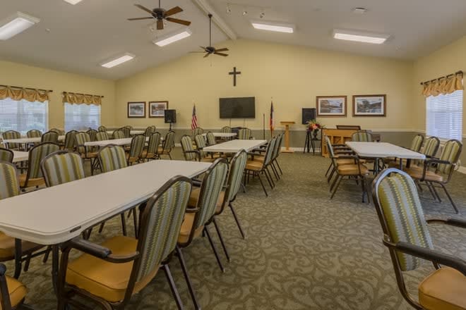 A spacious meeting room with multiple tables and chairs, featuring a cross on the wall, American flags, and a television.