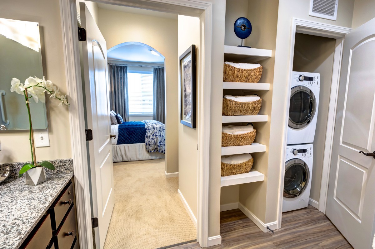 A cozy home interior featuring a hallway leading to a bedroom, with a laundry area equipped with stacked washer and dryer and shelves adorned with storage baskets.
