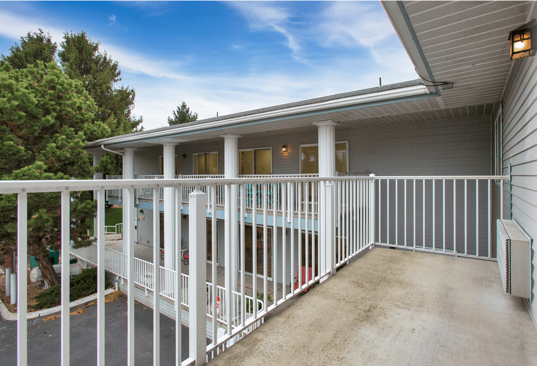 A view from a balcony with white railings overlooking a modern building and trees under a blue sky.