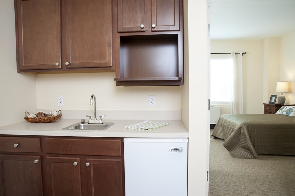 A compact kitchenette adjacent to a bedroom with wooden cabinets, a small sink, and a mini fridge.