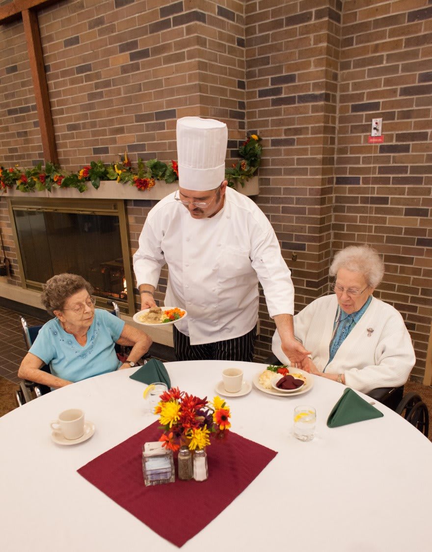 A chef serves meals to two seniors at a dining table.