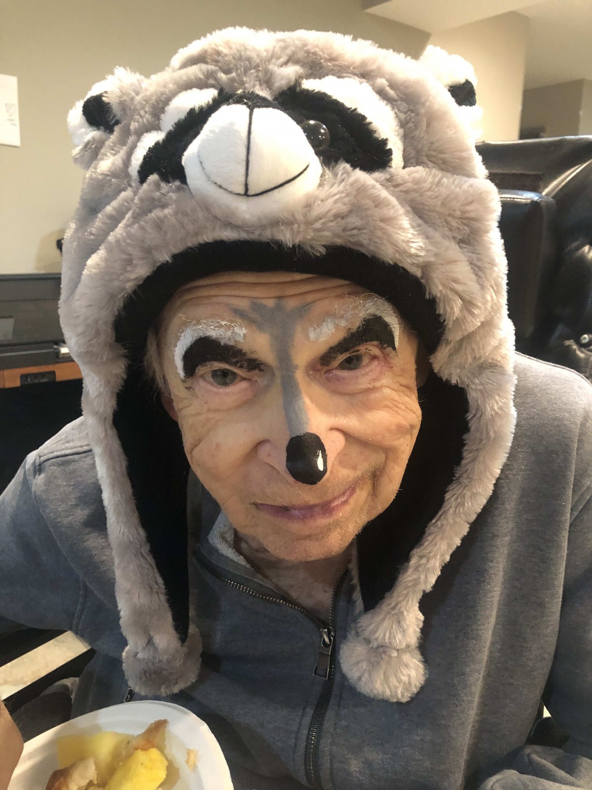An elderly person wearing a raccoon-themed hat and face paint.