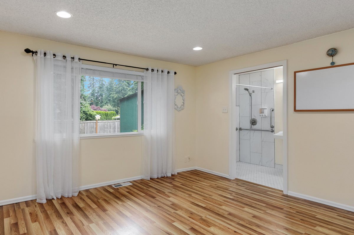 A bright bedroom with hardwood flooring, featuring a large window with sheer curtains and direct access to an adjacent bathroom.