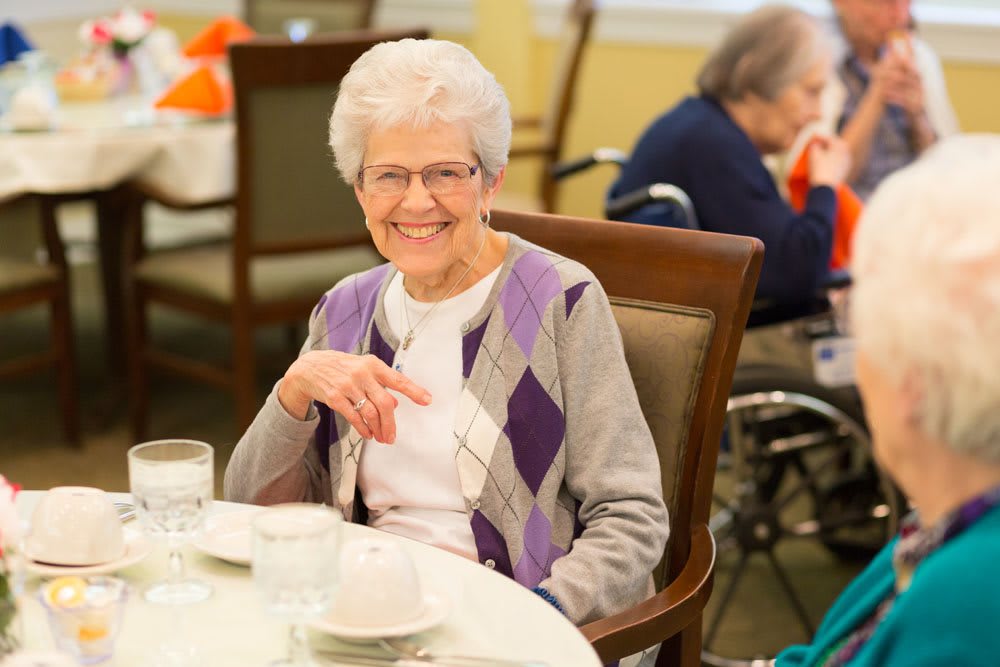 An elderly woman with glasses smiling while seated at a dining table indoors.