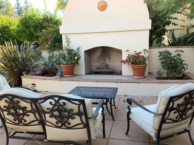 A cozy outdoor patio with a fireplace and comfortable seating surrounded by lush greenery.