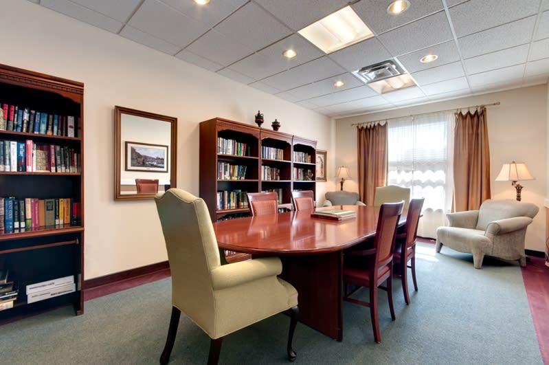 A well-lit conference room with a large wooden table surrounded by chairs, featuring a bookshelf and wall artwork.
